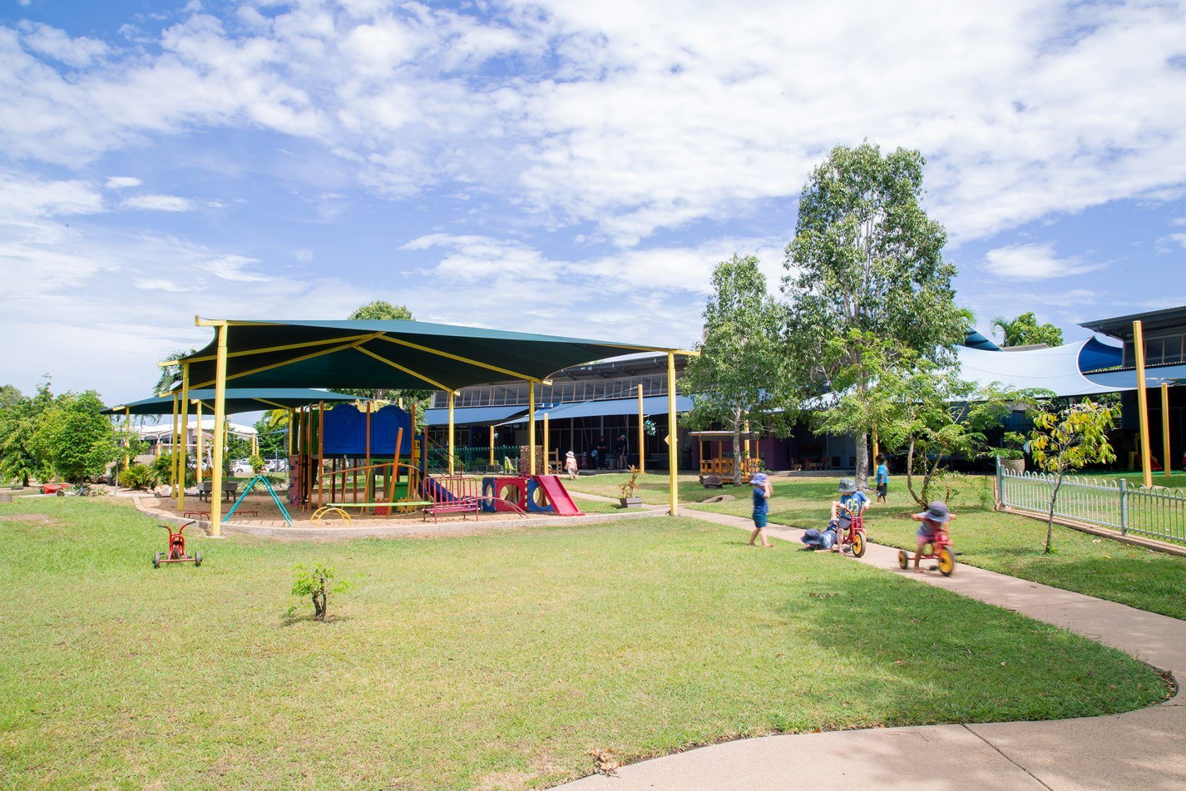 Children playing on a playground with a large shaded area and green lawn on a sunny day.