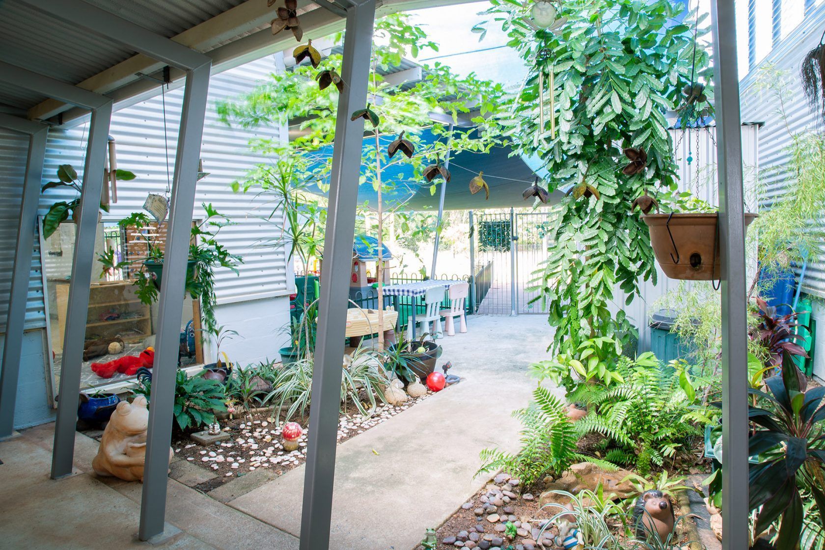 Courtyard with lush plants, vines, and hanging baskets under a corrugated metal roof. Pathway leads to a table and chairs.