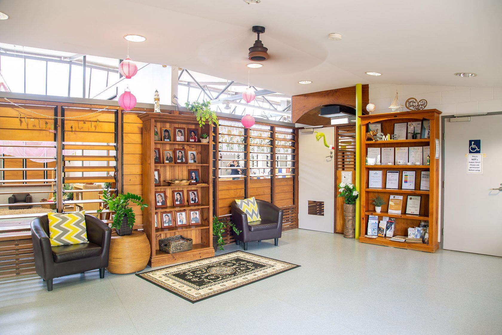 Cozy waiting area with wooden bookshelves, armchairs, and a decorative rug. Light-filled space with plants and framed photos.