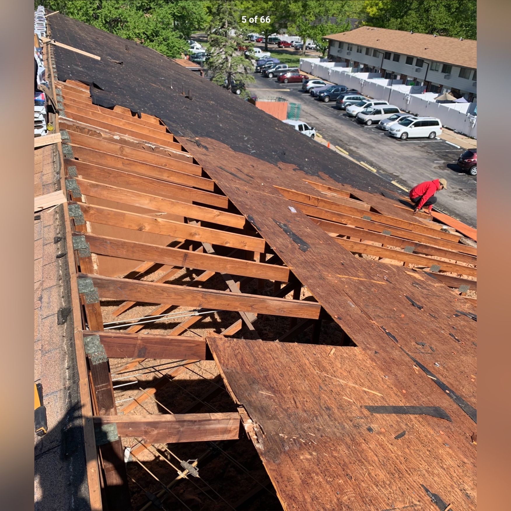 Roof partially stripped, revealing wooden structure with a worker in red, in a residential setting with parked cars.