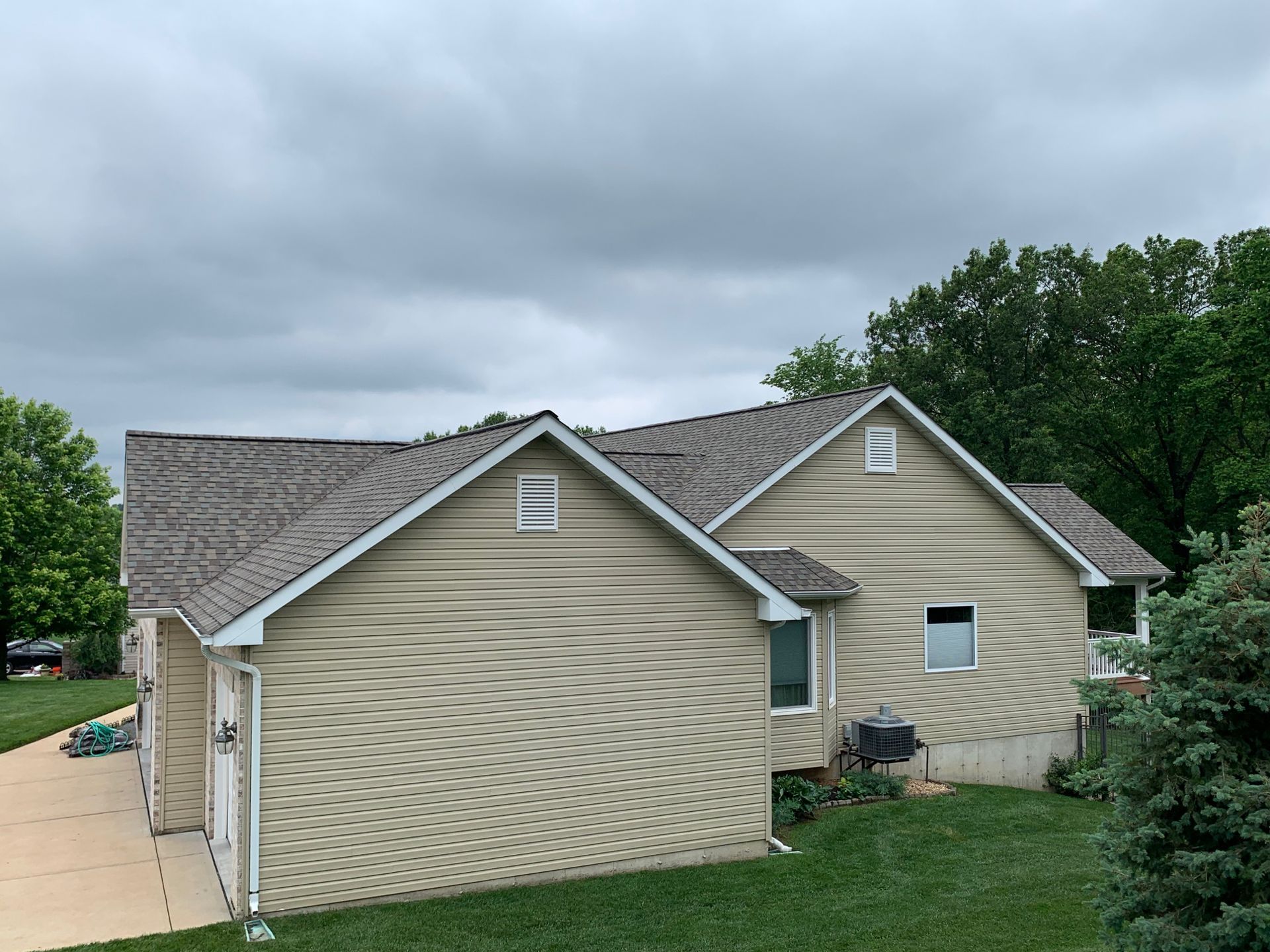 Beige house with gray roof under a cloudy sky. Green lawn and trees surround the house.