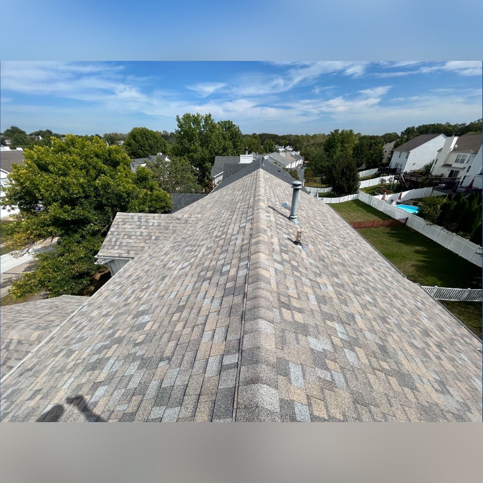 View from a rooftop showing a gray asphalt shingle roof with a metal chimney, surrounded by trees, houses, and a blue sky.