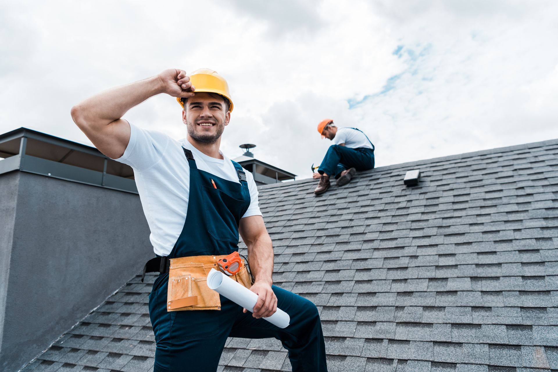 A happy roofer holding rolled paper while a coworker performs residential roofing services. A happy roofer holding rolled paper while a coworker performs residential roofing services.