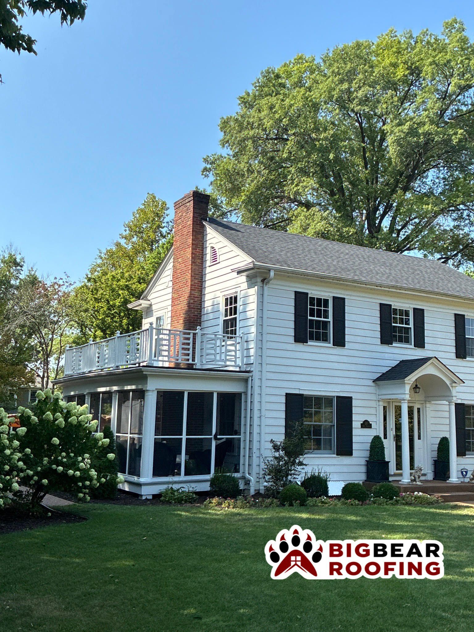 White house with black shutters, screened porch, chimney, and green yard under a blue sky.