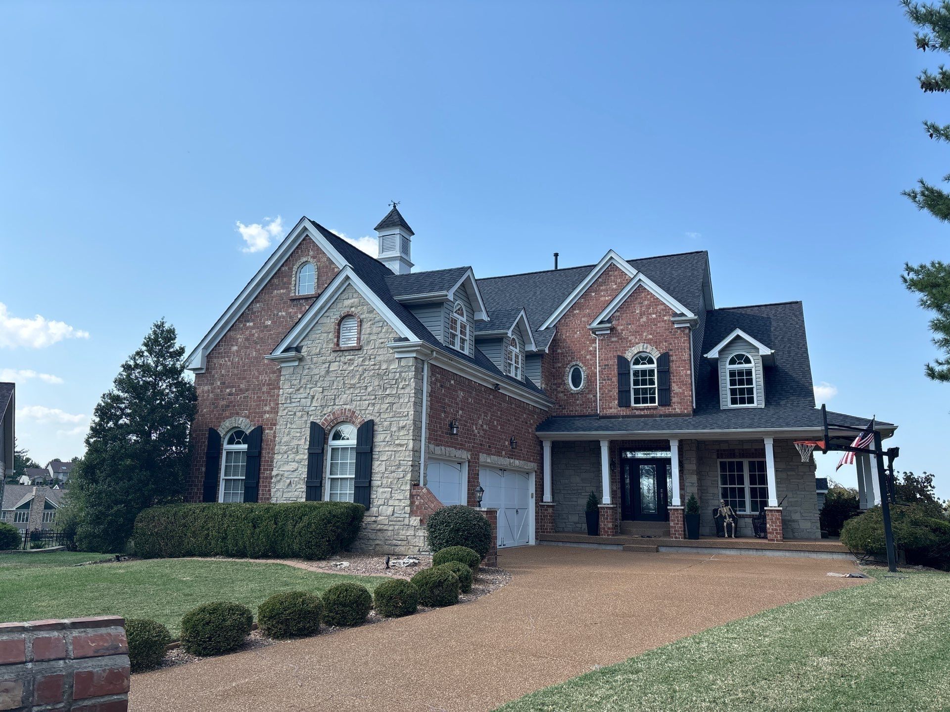 Large brick and stone house with a gravel driveway and green lawn under a blue sky.