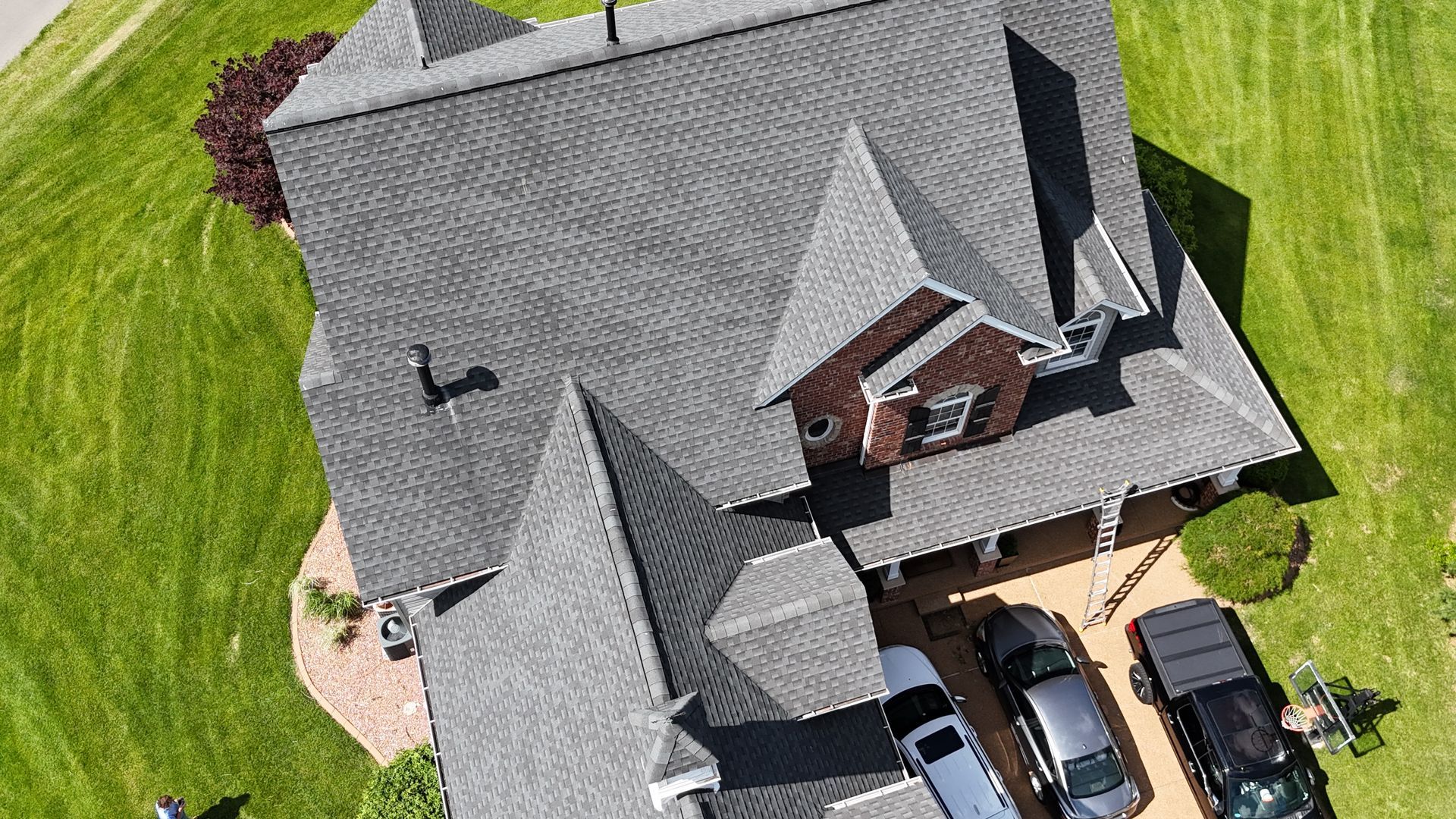 Aerial view of a gray-roofed house with cars in the driveway, surrounded by green grass.
