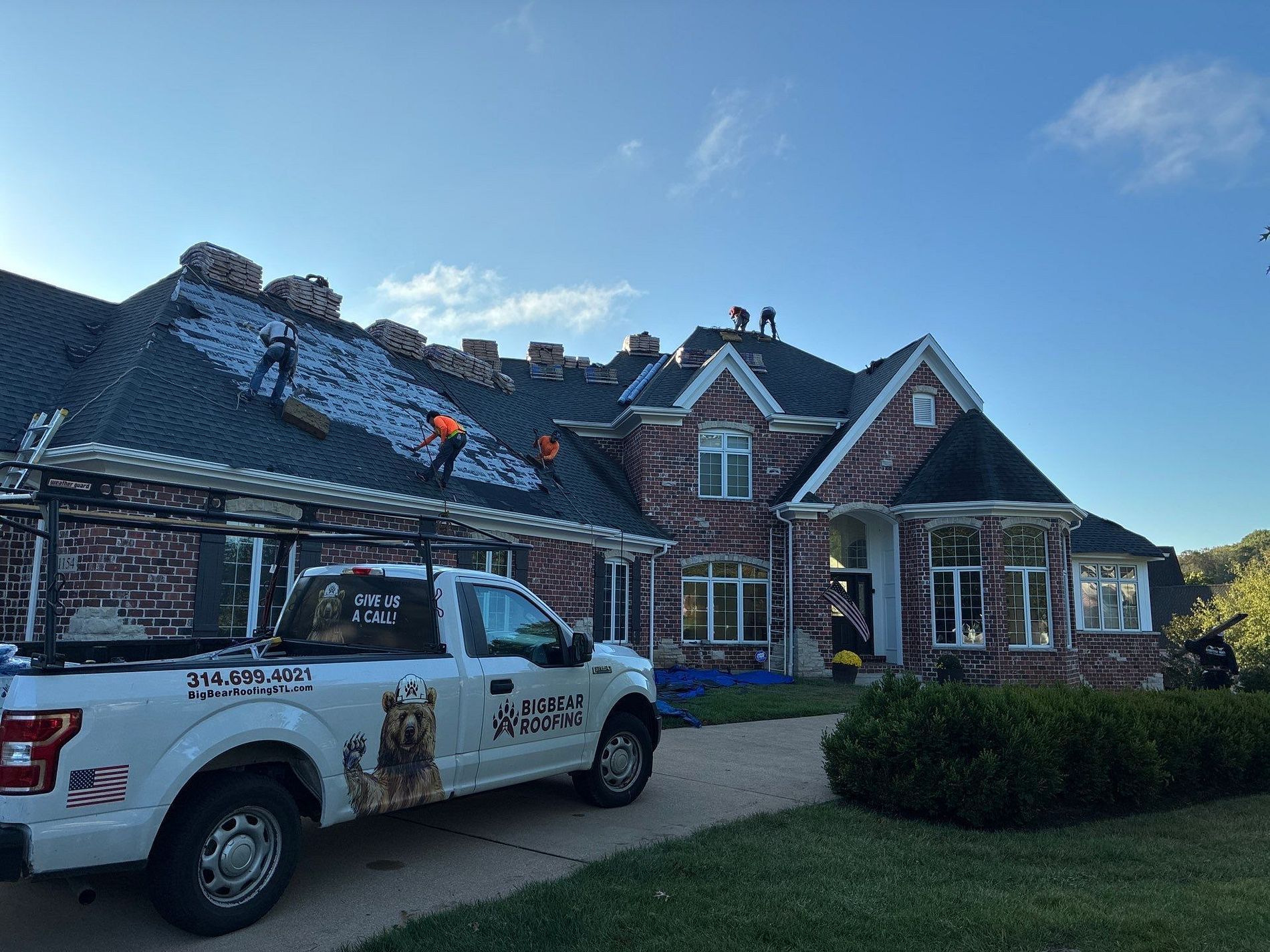 Roofers on a house with a red brick facade, working near a white truck with a bear logo, blue tarp on the ground.