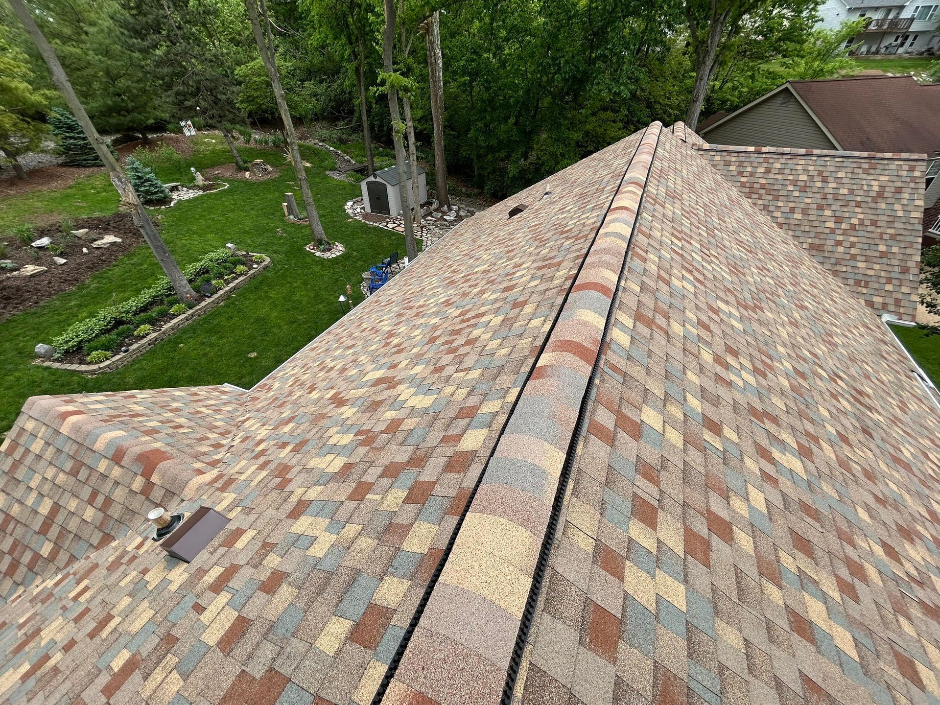 Overhead view of a roof with colorful shingles and a central ridge, surrounded by trees and a lawn.