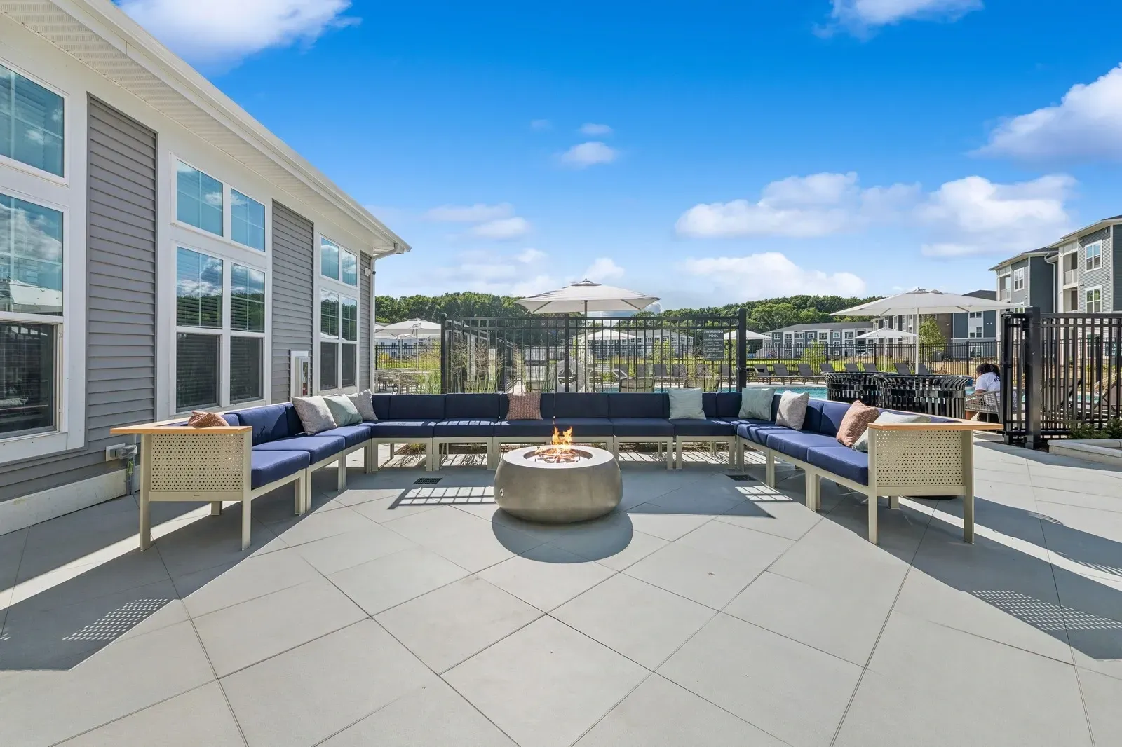 Outdoor amenity area with a circular fire pit, cushioned seating, umbrellas, and a person nearby.