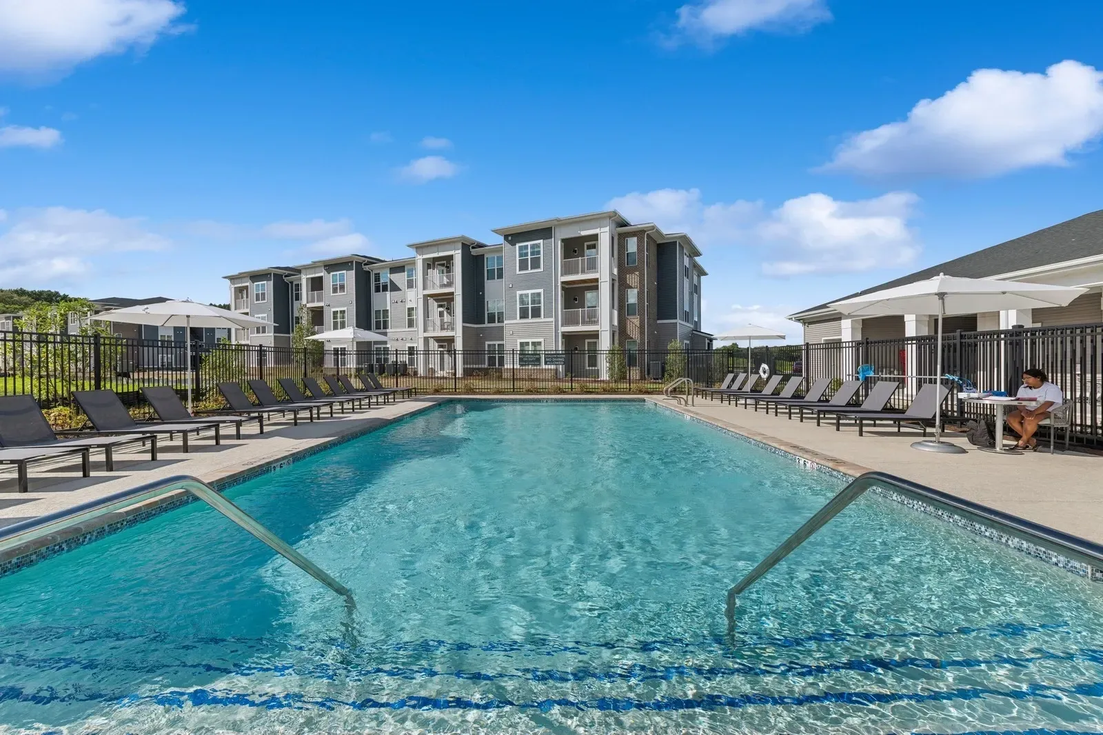 Pool area at an apartment community with lounge chairs, umbrellas, and a person seated at a table.