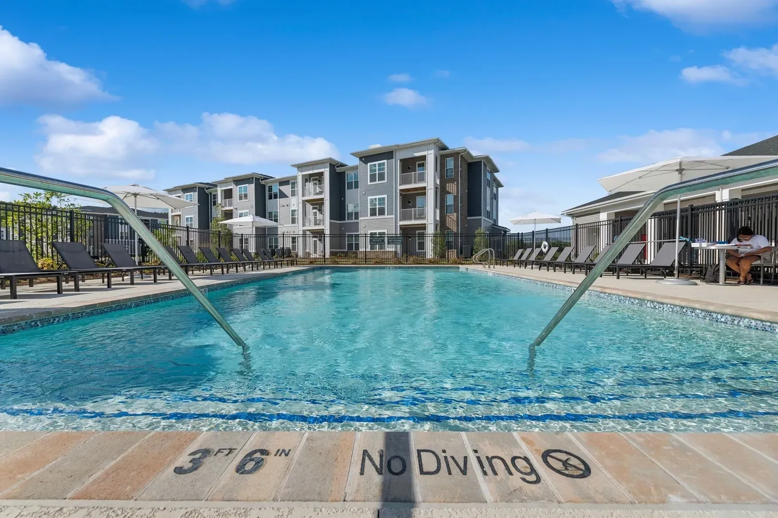 Person sitting by a modern apartment community pool with lounge chairs and umbrellas.