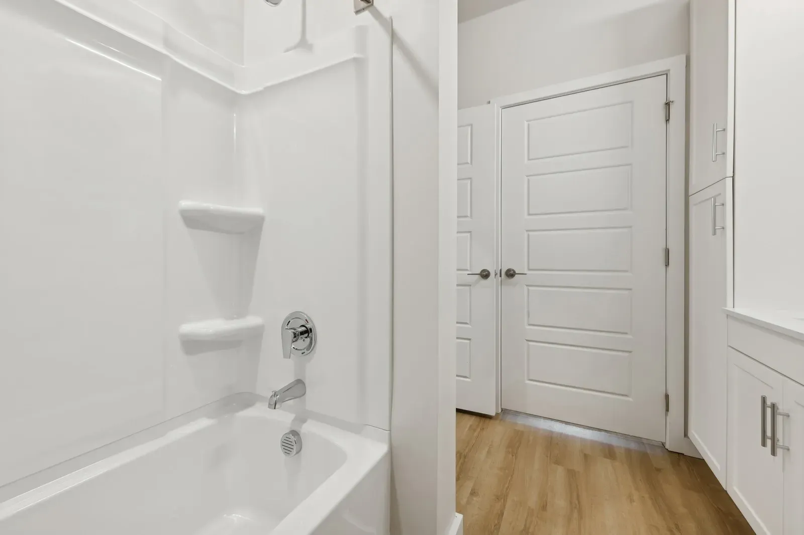 White bathroom with tub/shower enclosure, chrome fixtures, and white cabinets; light wood flooring.