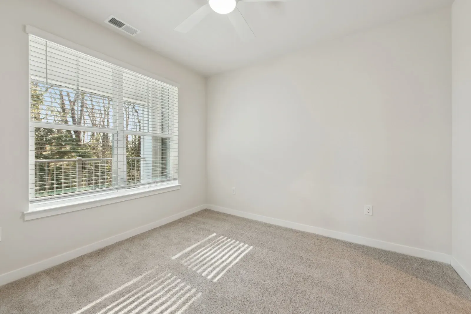 Empty bedroom with a large window, white blinds, ceiling fan, and beige carpet.