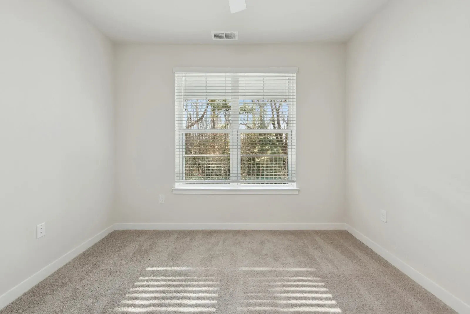 Small empty bedroom with light walls, beige carpeting, and a window with horizontal blinds.