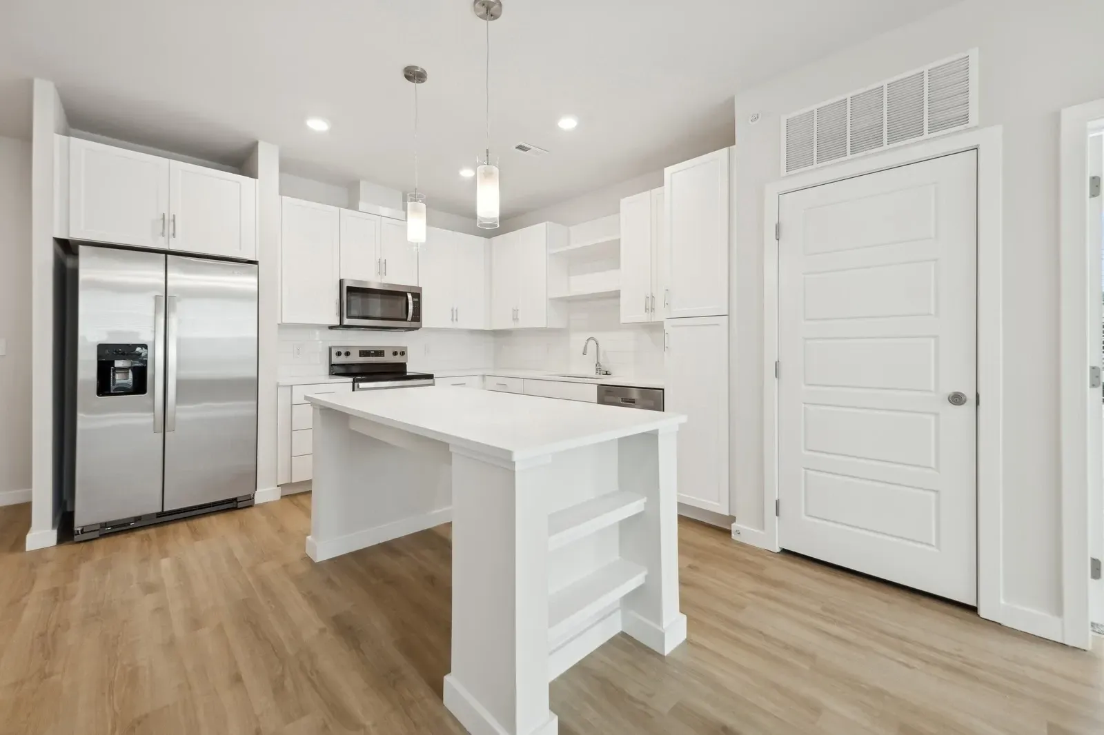 Bright white kitchen in an apartment featuring an island, stainless steel appliances, and light wood flooring.