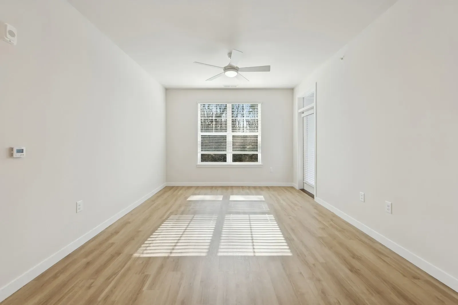 Empty living room with wood floors, window blinds, and a ceiling fan.