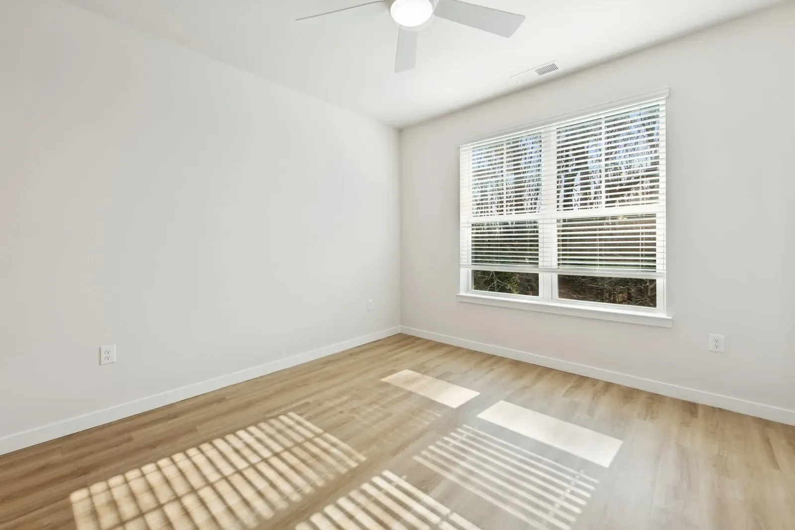 Empty bedroom with white walls, a large window with blinds, wood-look flooring, and a ceiling fan.