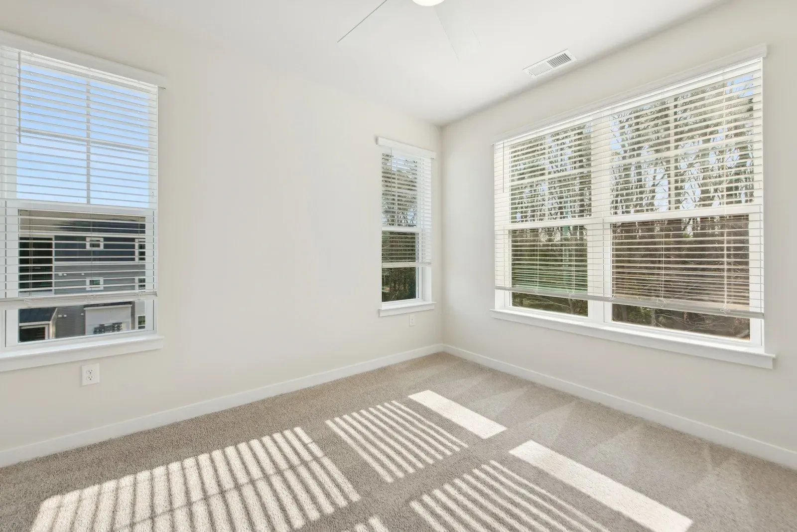 Bright empty bedroom with beige carpet, white walls, and large windows with horizontal blinds.