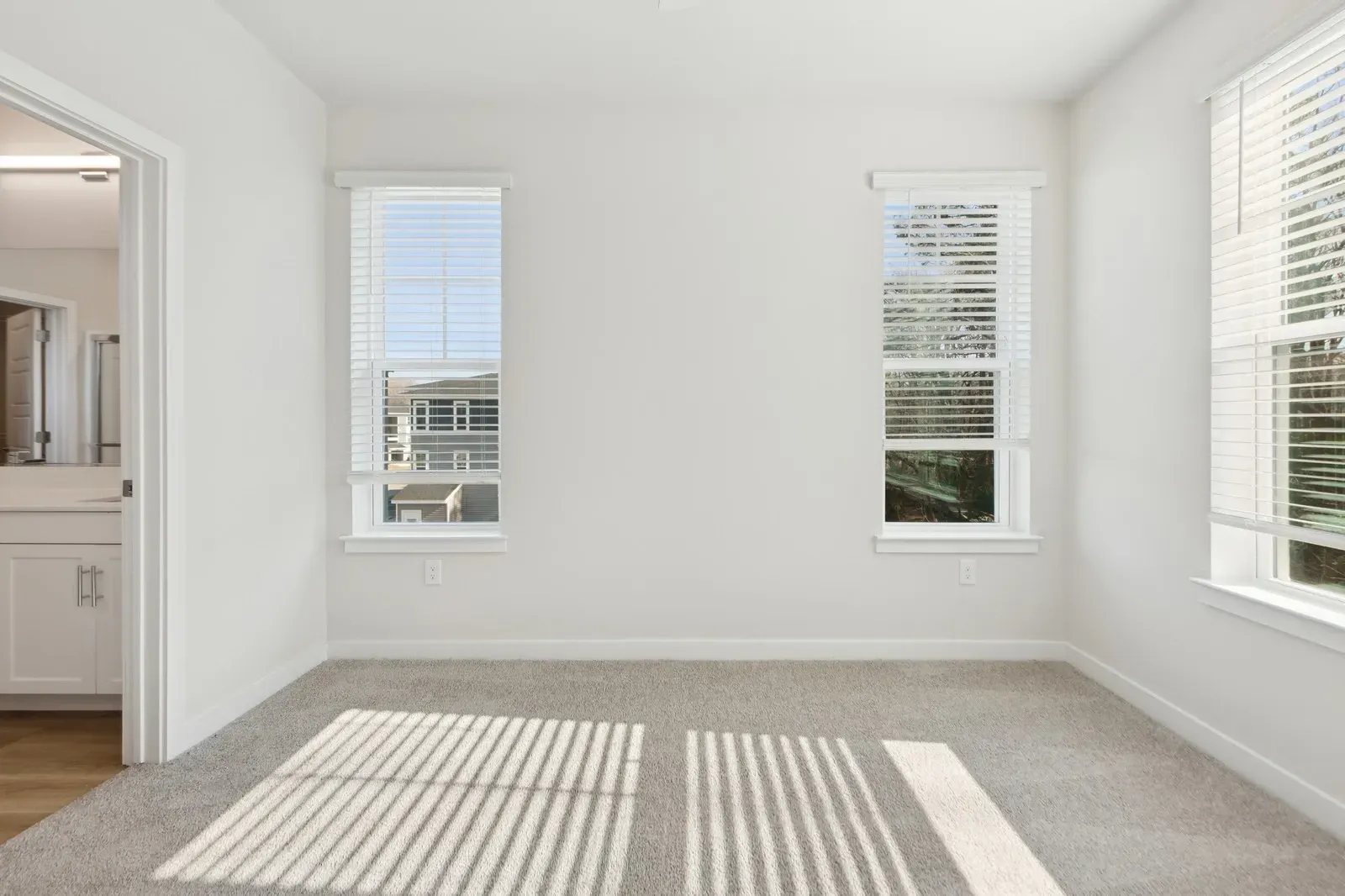Bright empty bedroom with two windows, white walls, beige carpet, and an open doorway to a bathroom.
