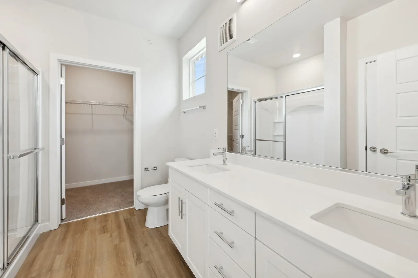 Modern white bathroom with double vanity, large mirror, shower, and walk-in closet.