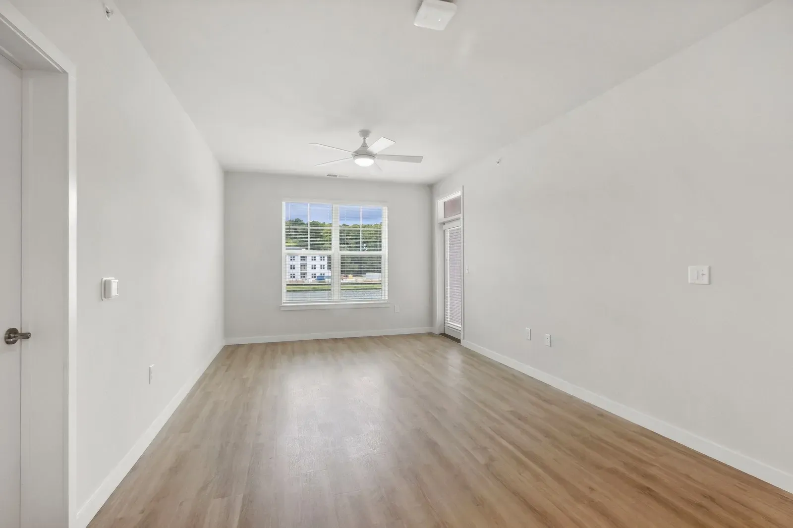 Empty apartment living room with large window, balcony door, ceiling fan, and light wood floors.