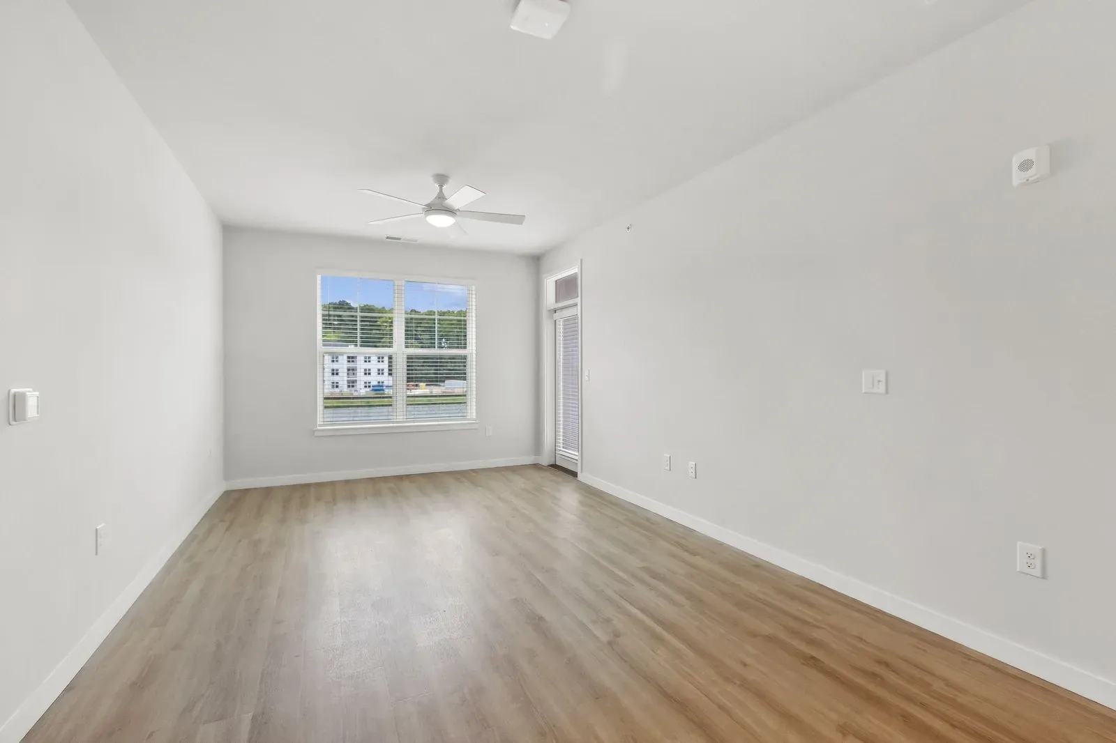 Empty living room with wood-like flooring, white walls, ceiling fan, and a large window