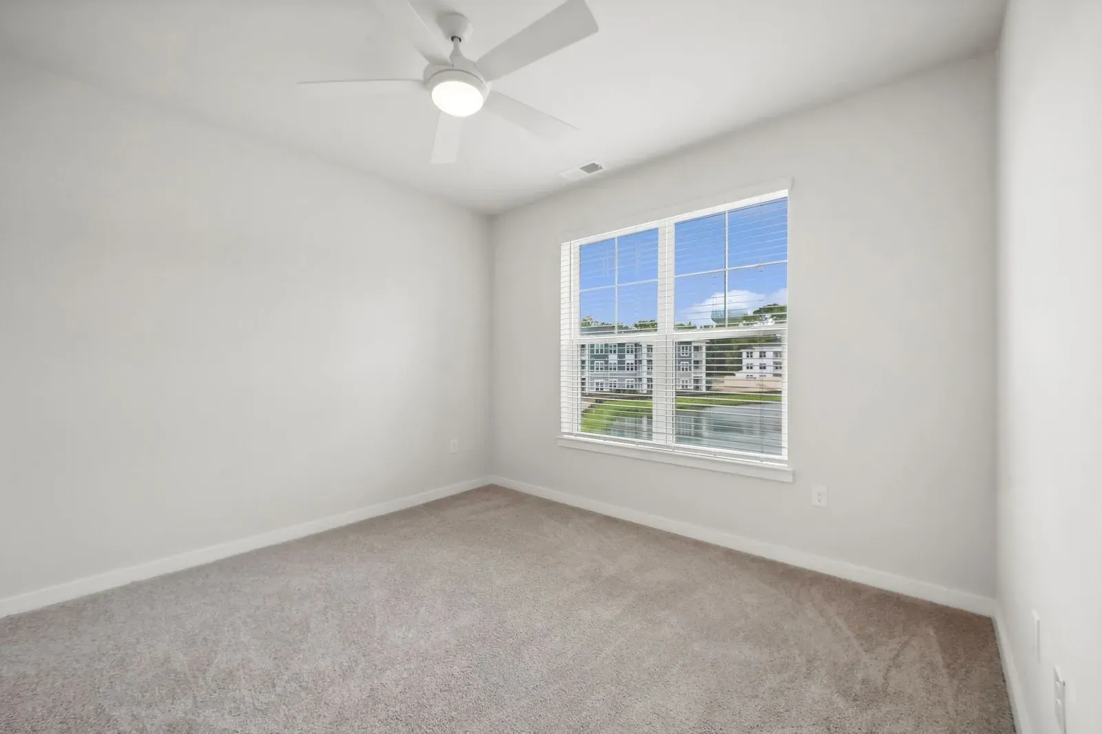 Empty bedroom with white walls, beige carpet, ceiling fan, and a window with blinds.