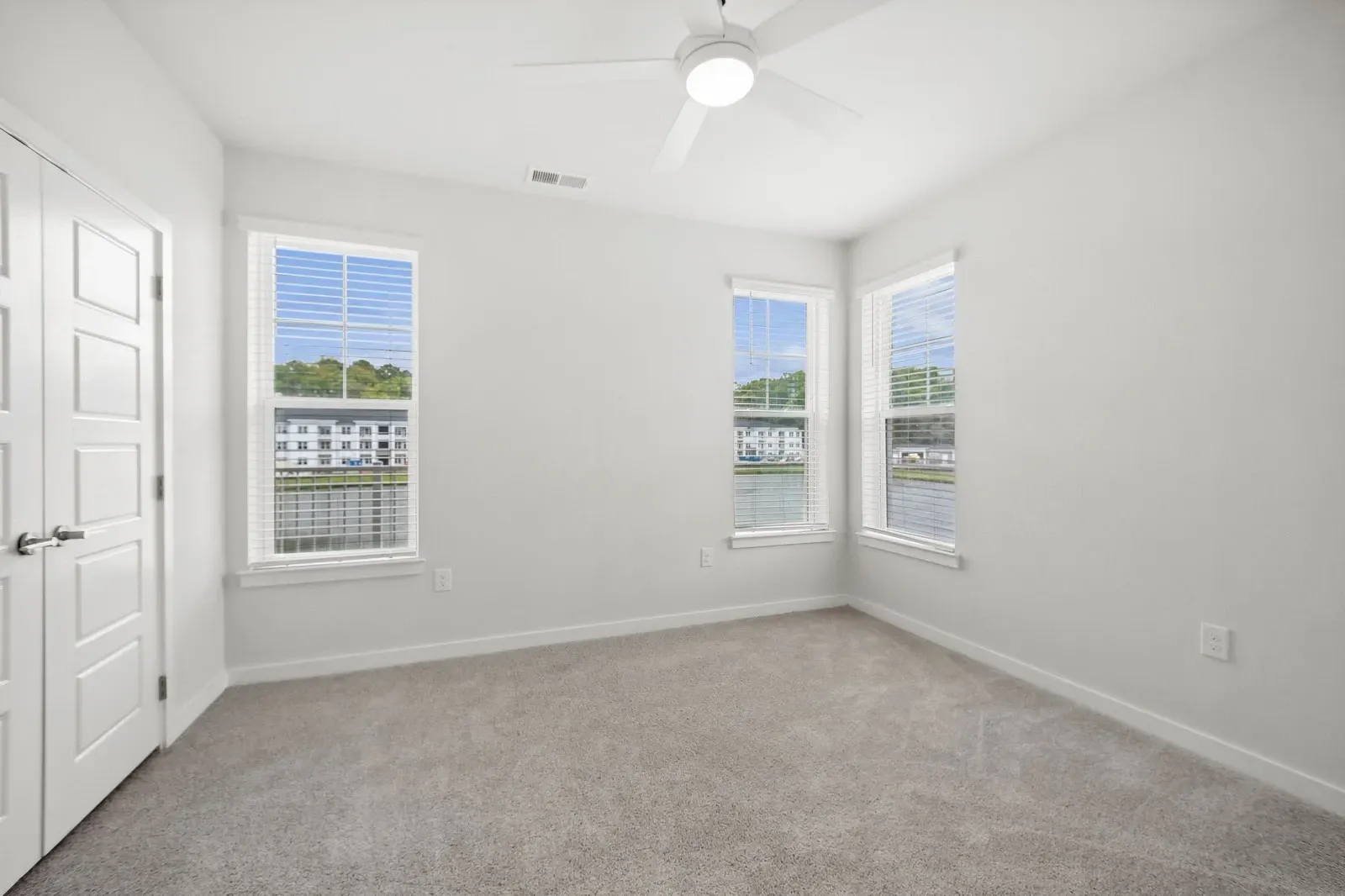 Bright, unfurnished bedroom with white walls, beige carpet, and a ceiling fan, featuring large windows.