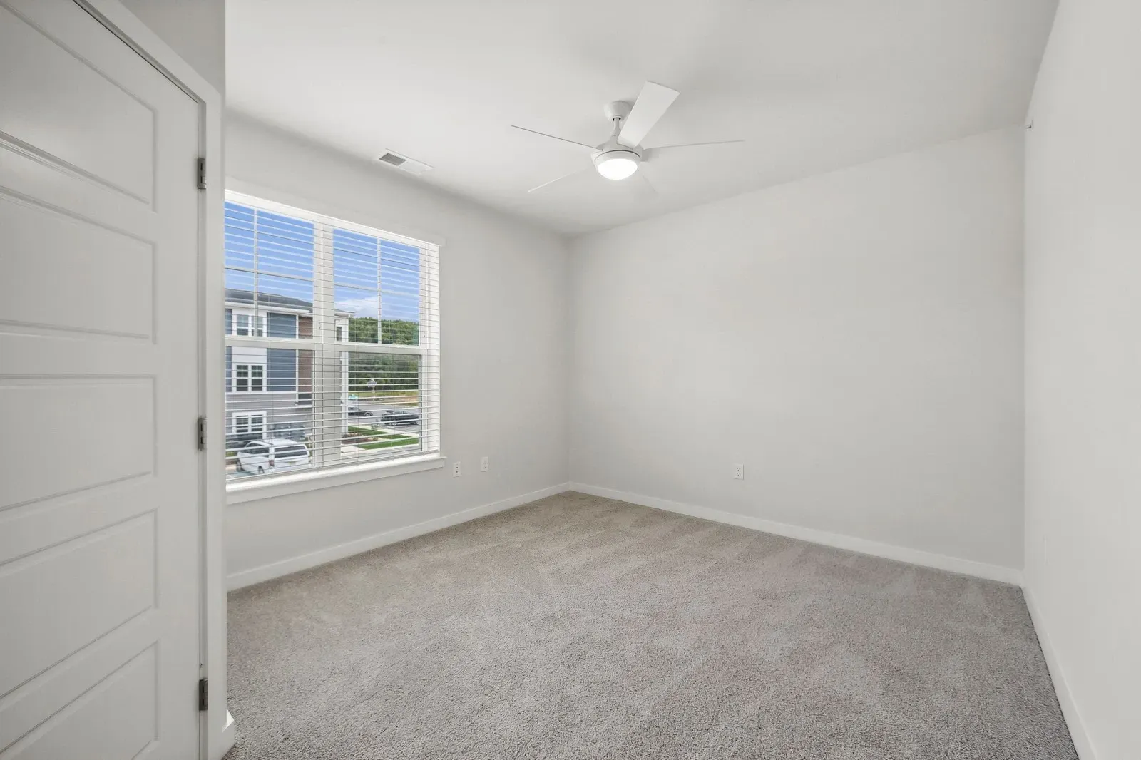 Bright empty bedroom with white walls, beige carpet, a large window with white blinds, and a ceiling fan.