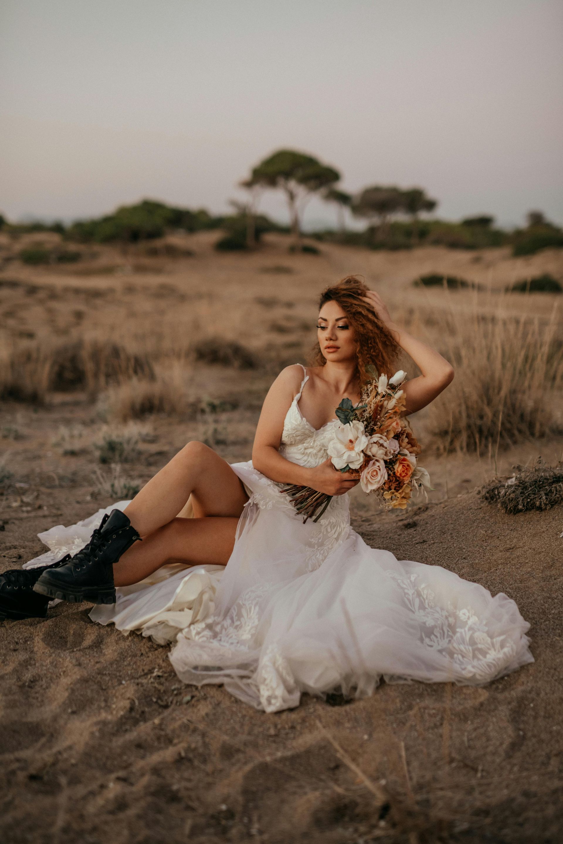 A woman in a wedding dress is sitting on the sand holding a bouquet of flowers.