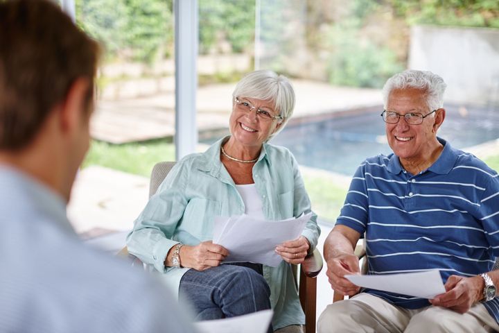 An elderly couple is sitting in chairs looking at papers while a man talks to them.