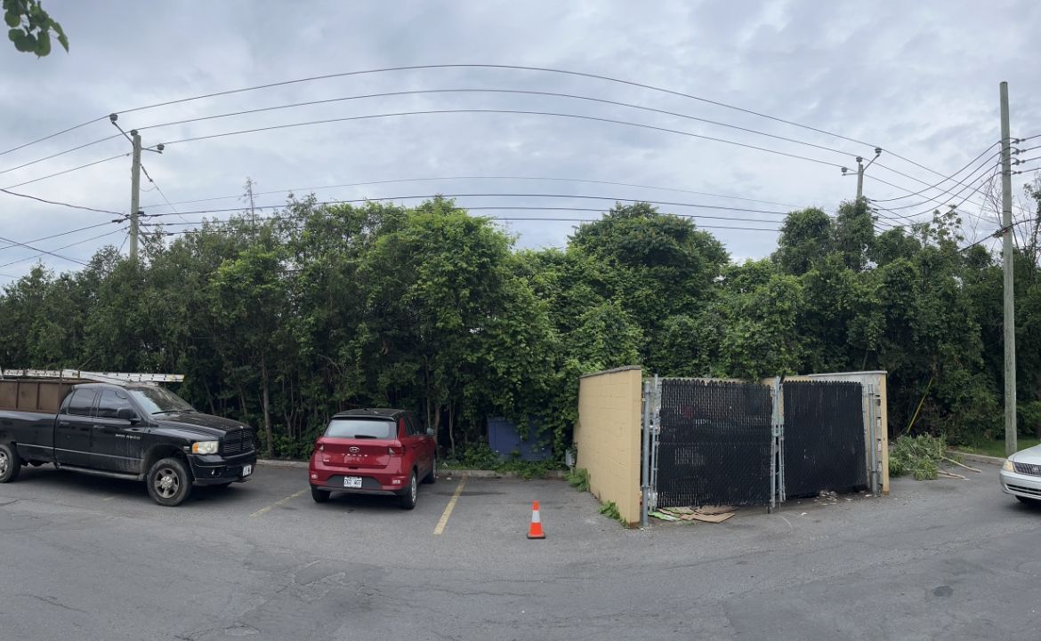 Parking avec voitures, arbres, poteaux électriques et lignes électriques aériennes. Ciel nuageux.
