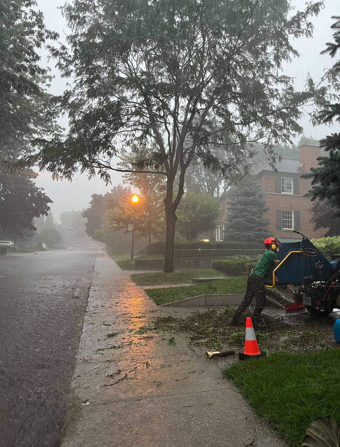 Une personne utilise un broyeur de branches dans une rue résidentielle détrempée. Pluie, arbres et maisons sont visibles en arrière-plan.