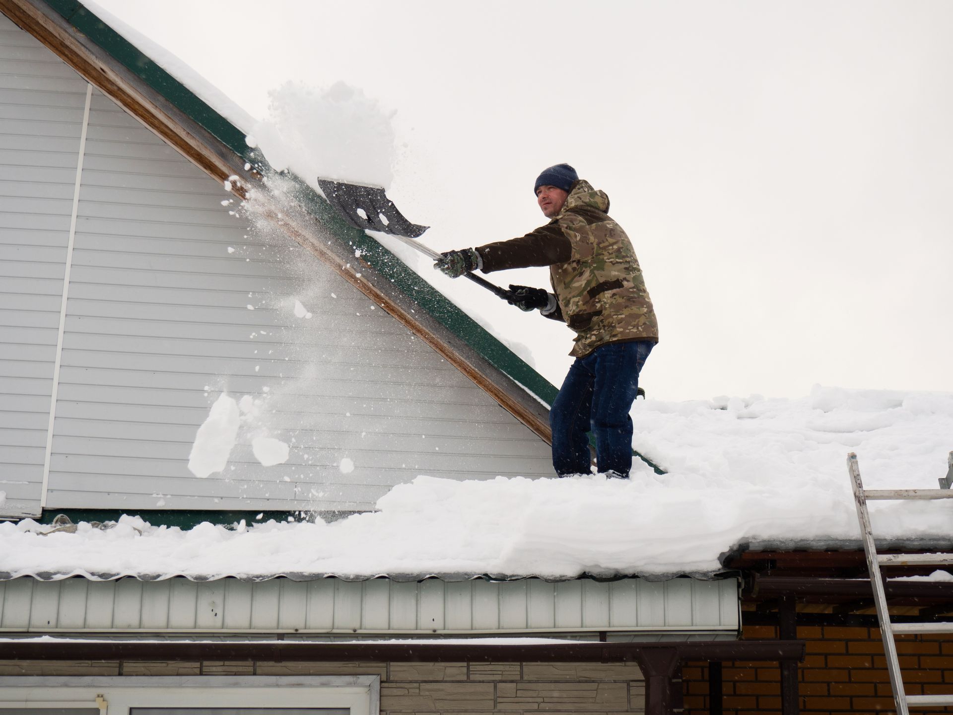 Un homme déneige un toit à l'aide d'un râteau. Journée nuageuse.