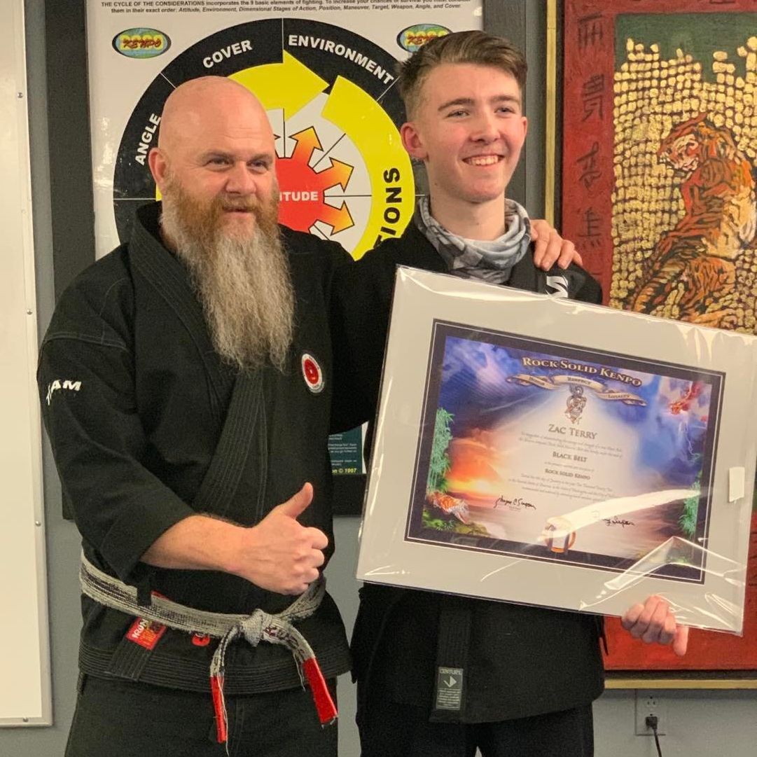 A man with a beard is standing next to another man holding a framed picture