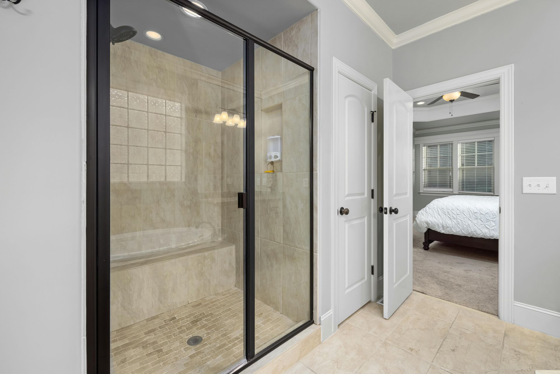 Bathroom with a glass shower, two white doors, and a doorway to a bedroom with a bed.