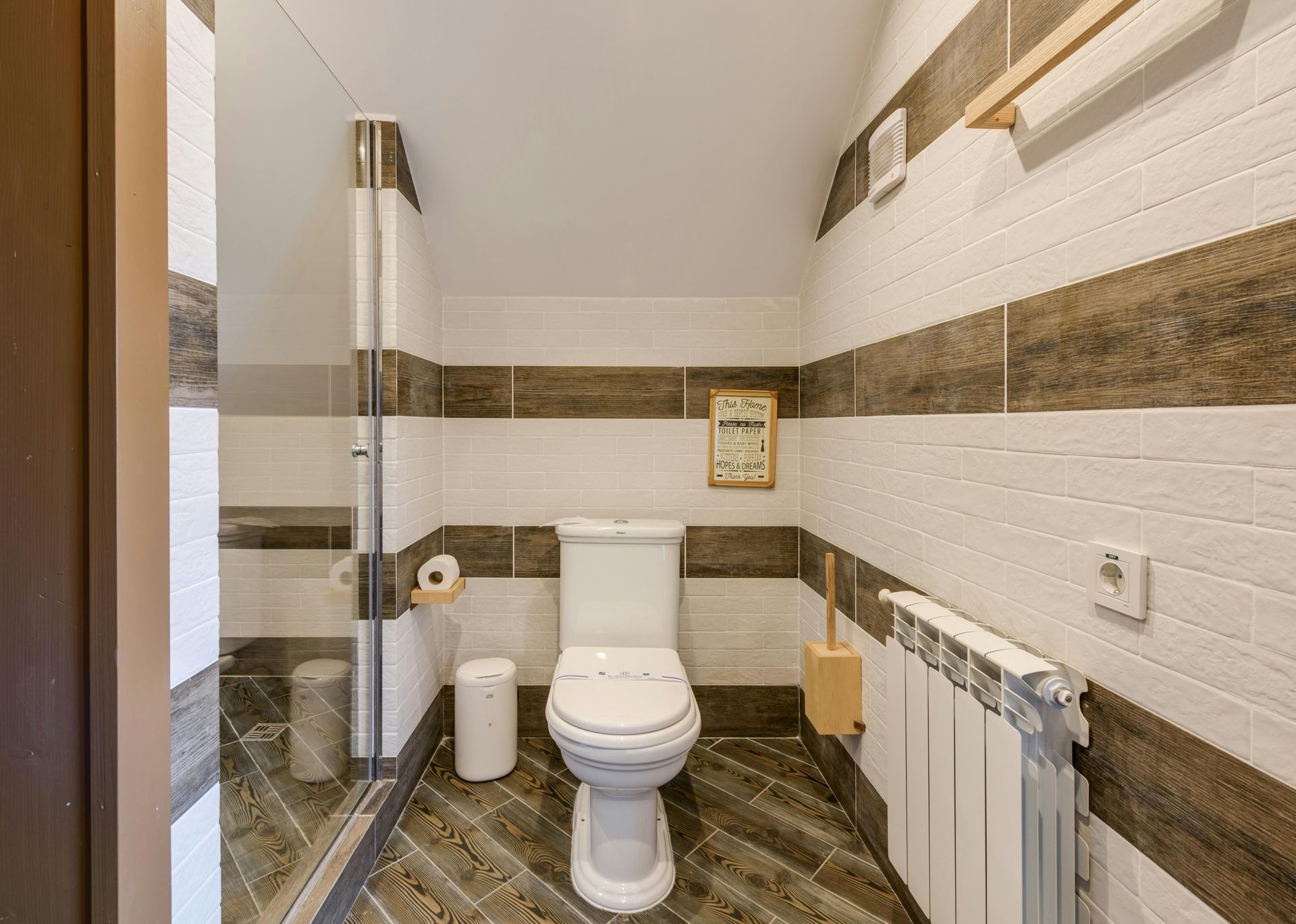 Small bathroom with a toilet, glass shower, and radiator, decorated with horizontal brown and white tiles.