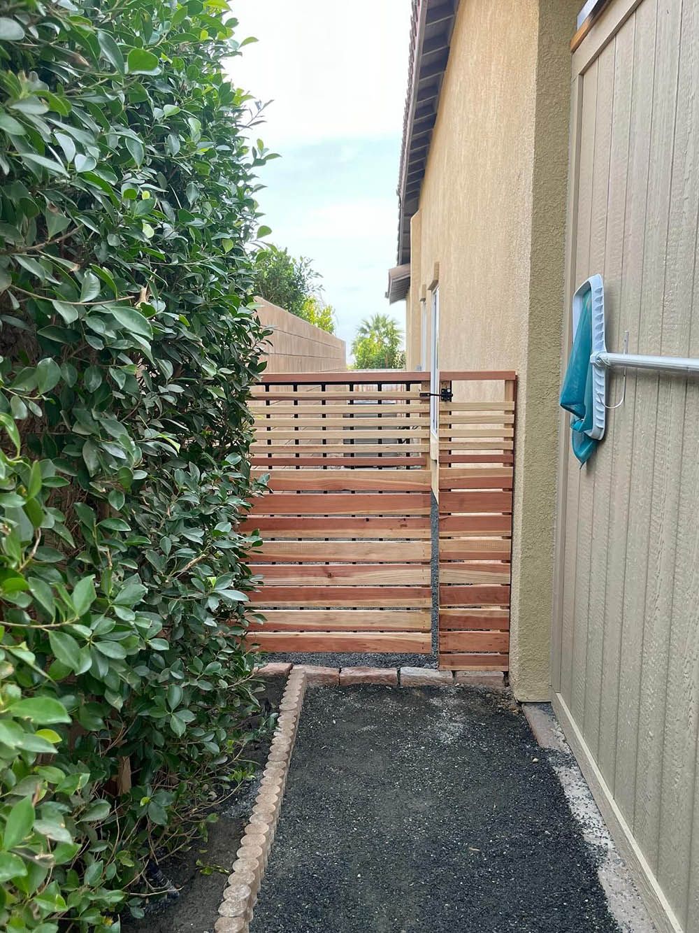 A wooden gate is between a house and a hedge.