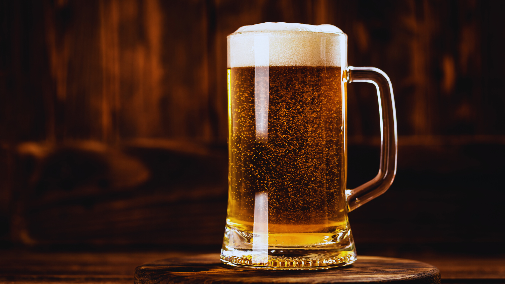 Glass of beer with a frothy head, on a wooden surface against a dark wood background.