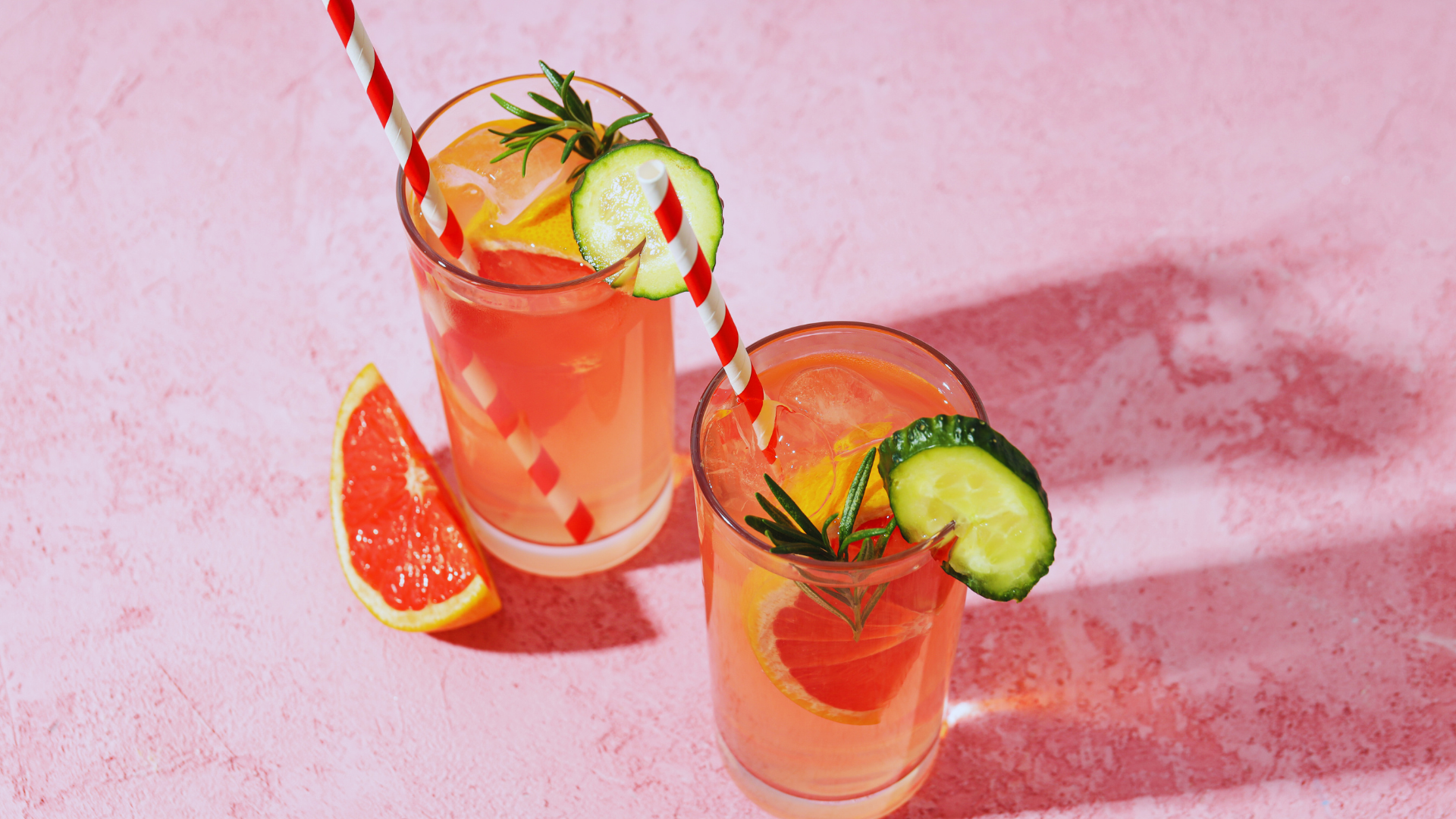 Two glasses of pink cocktails with grapefruit, cucumber, and rosemary garnishes; pink background.