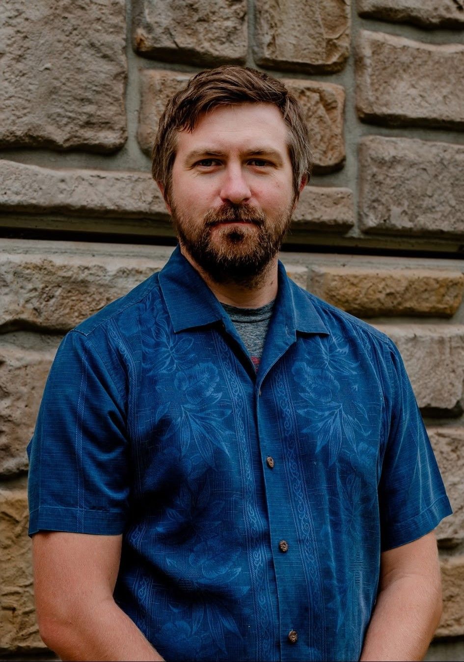 Man in a blue shirt standing against a stone wall, looking at the camera.