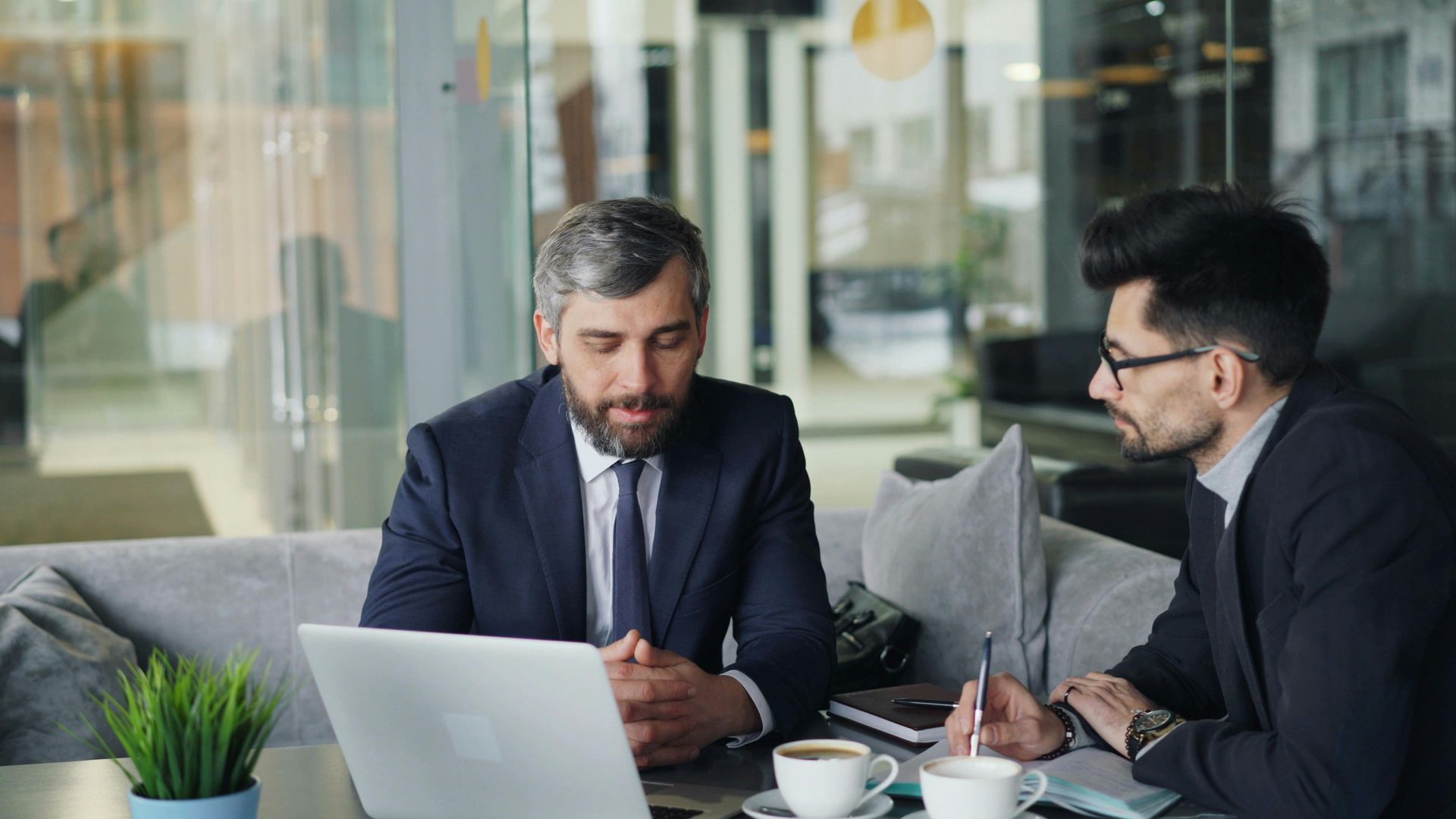 Two professionals in suits discussing documents at a café table with a laptop and coffee.