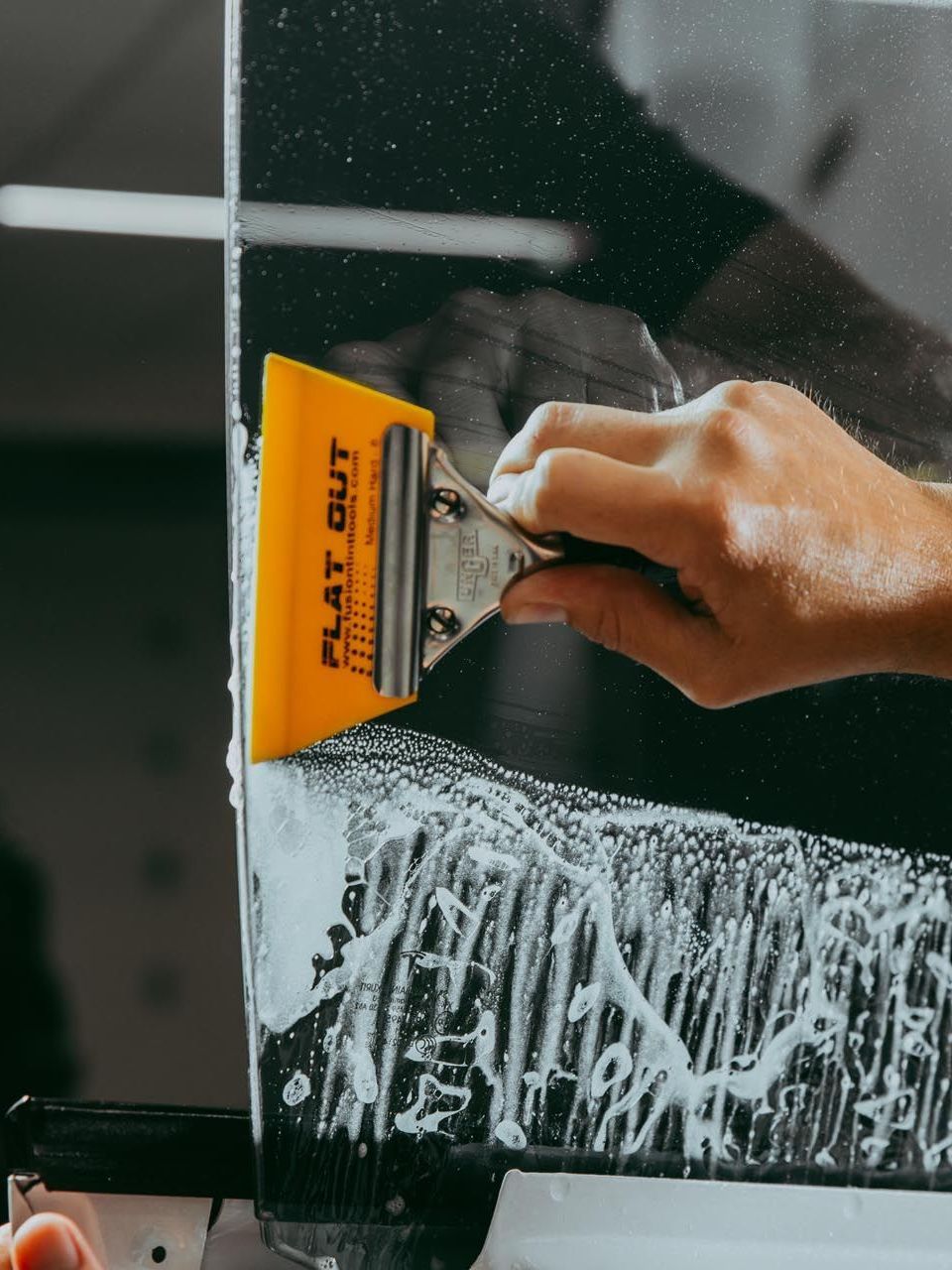 Person using a yellow squeegee to smooth a layer of soapy solution on black surface, likely applying tint film.