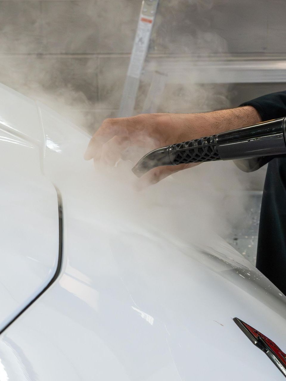 A person uses a steam cleaner on a white car, releasing a cloud of steam. A ladder is in the background.