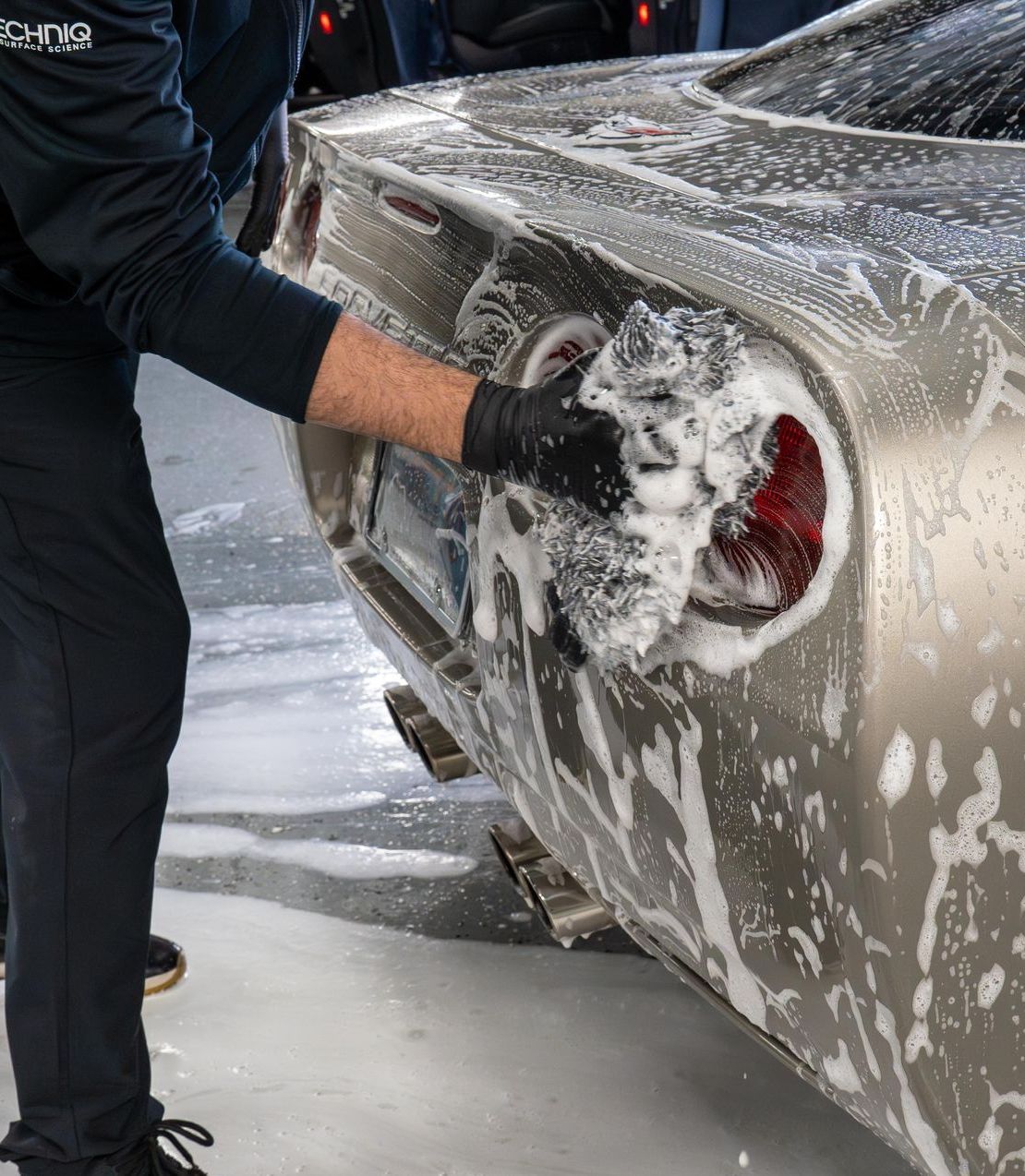 Person washing a silver sports car's rear with a soapy sponge. Black gloves and pants are visible.