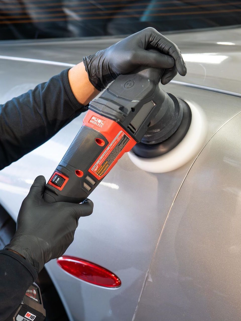 A person in black gloves polishing a silver car with a red and black buffer in a well-lit garage.