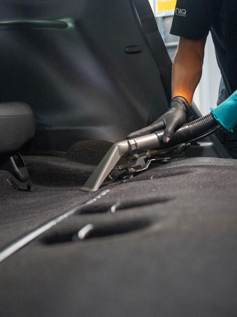 A person in black gloves vacuums the dark car carpet with a handheld tool inside a vehicle.
