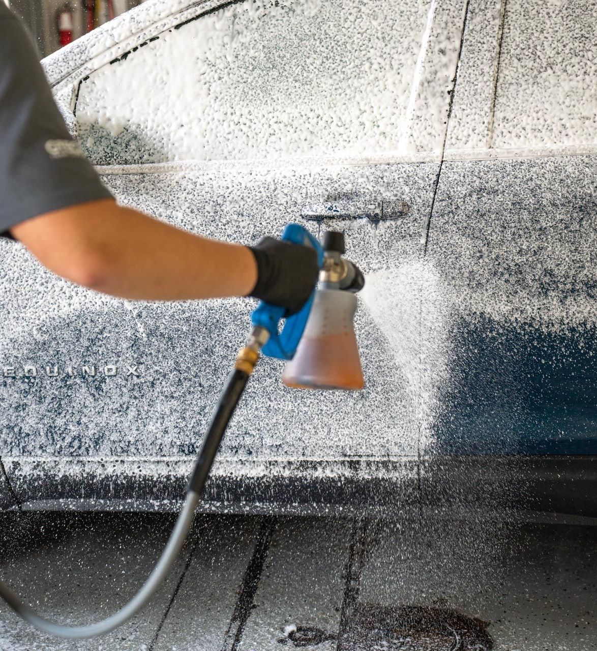 Person sprays foamy car wash solution onto a blue car door using a foam cannon in a garage setting.