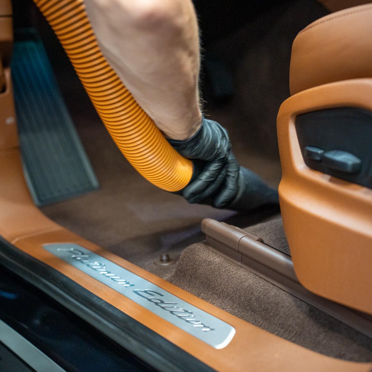 Person vacuuming the brown carpet of a car interior. The person wears black gloves and holds an orange hose.