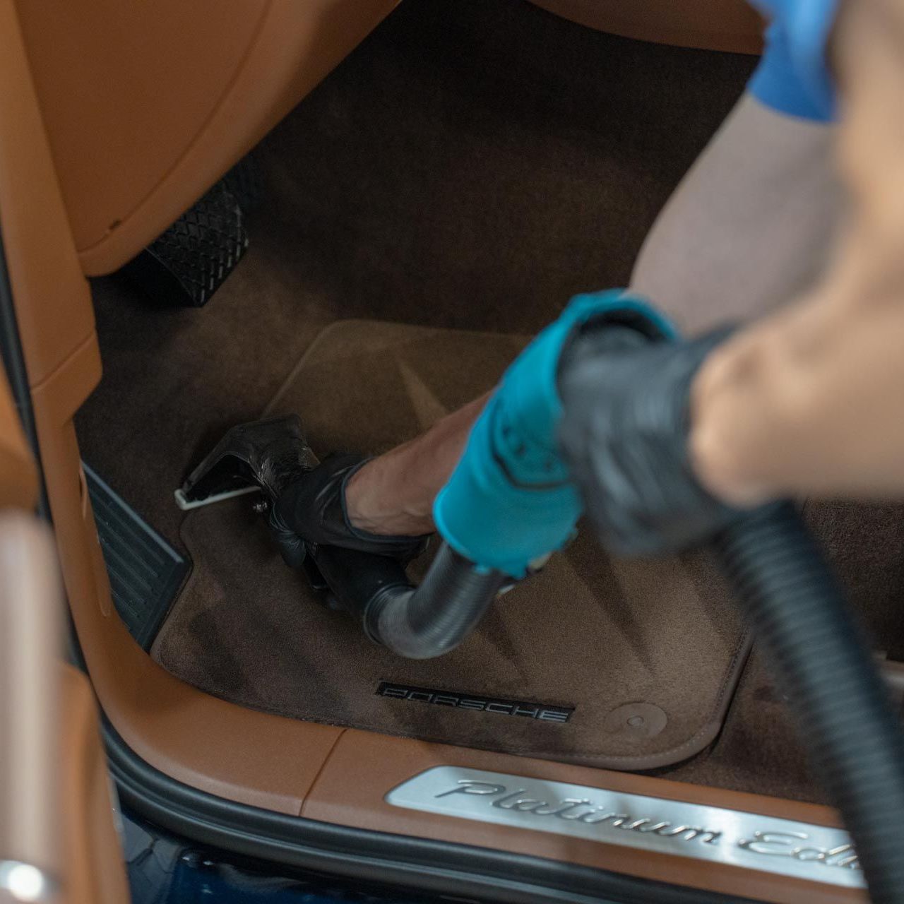 A person wearing a black glove vacuums the brown carpet inside a car, near the